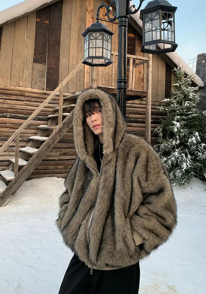 Person wearing a large brown fur coat in a snowy landscape with a wooden cabin.